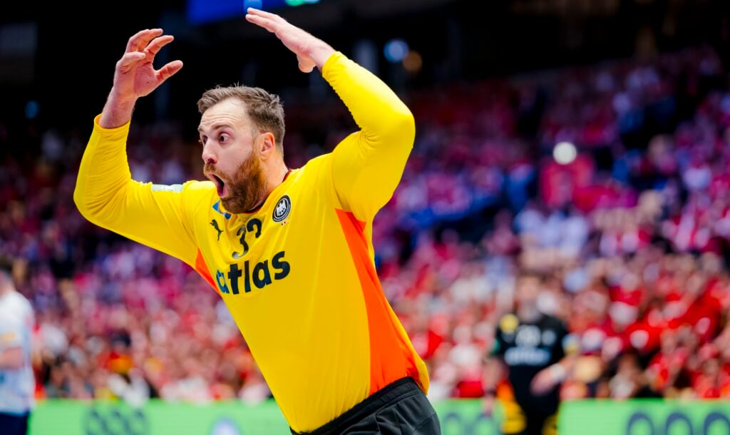 Ein Handballtorwart in einem gelben Trikot mit einem Atlas auf der Vorderseite hebt die Arme und sieht während eines Spiels überrascht oder aufgeregt aus, mit einer verschwommenen Menge und Spielern im Hintergrund.