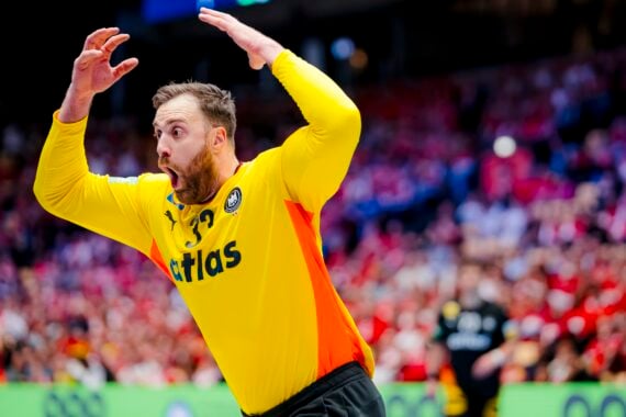 Ein Handballtorwart in einem gelben Trikot mit einem Atlas auf der Vorderseite hebt die Arme und sieht während eines Spiels überrascht oder aufgeregt aus, mit einer verschwommenen Menge und Spielern im Hintergrund.