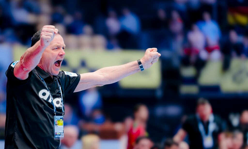 Ein Mann in einem schwarzen Hemd reckt begeistert beide Fäuste in die Höhe und jubelt bei einem Sportereignis. Er trägt ein Schlüsselband und steht vor einer verschwommenen Menschenmenge im Hintergrund.