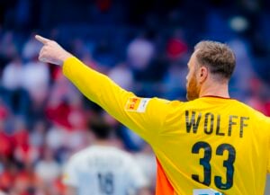 Ein Handballtorwart, der ein gelbes Trikot mit dem Namen Wolff und der Nummer 33 trägt, gestikuliert und zeigt mit seiner linken Hand; im Hintergrund sind unscharfe Spieler und Zuschauer zu sehen.