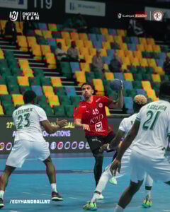 Ein männlicher Handballspieler in einem roten Trikot springt, um den Ball zu werfen, während drei gegnerische Spieler in weißen Trikots ihn auf einem Hallenplatz mit grünen und gelben Sitzen im Hintergrund verteidigen.