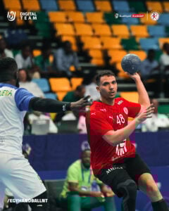 Ein männlicher Handballspieler in einem roten Trikot mit der Nummer 48 bereitet sich auf den Wurf vor, während ein Verteidiger in einem hellblauen Trikot ihm gegenübersteht. Die Zuschauer sitzen auf der Tribüne hinter ihnen.