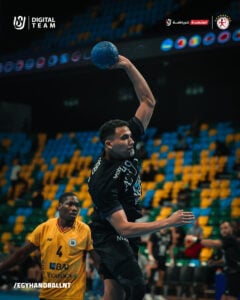 Ein männlicher Handballspieler in einem schwarzen Trikot springt, um den Ball zu werfen, während ein Verteidiger in einem gelben Trikot zusieht. Das Spiel findet in einem Hallenstadion mit bunten leeren Sitzen statt. Mannschafts- und Sponsorenlogos erscheinen auf dem Bild.