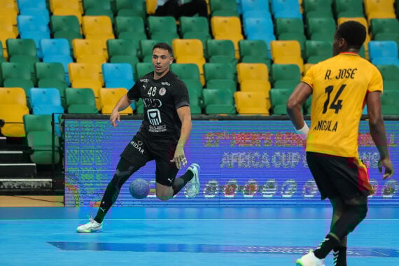 Ein Handballspieler in einem schwarzen Trikot bereitet sich auf den Wurf vor, während ein anderer Spieler in einem gelben Trikot mit R. José und 14 auf dem Rücken in der Nähe steht. Im Hintergrund sind leere grüne und gelbe Plätze zu sehen.