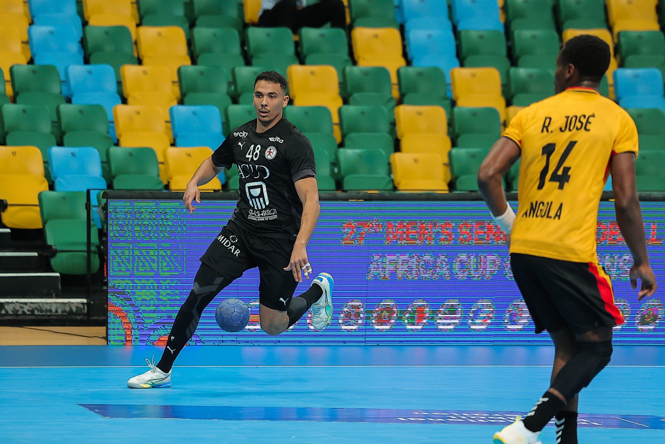 Ein Handballspieler in einem schwarzen Trikot bereitet sich auf den Wurf vor, während ein anderer Spieler in einem gelben Trikot mit R. José und 14 auf dem Rücken in der Nähe steht. Im Hintergrund sind leere grüne und gelbe Plätze zu sehen.