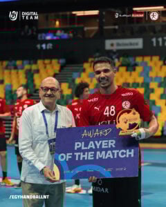 Ein lächelnder Handballspieler in einem roten Trikot hält eine Auszeichnung als "Spieler des Spiels" in der Hand und posiert neben einem älteren Mann in einem weißen Hemd auf einem Hallenplatz, mit anderen Spielern und leeren Sitzen im Hintergrund.