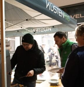 Zwei Personen unter einem Zelt servieren Essen von einem Büffettisch im Innenbereich. Einer trägt eine schwarze Jacke und einen Hut, der andere eine grüne Weste. Die Teller stapeln sich auf dem Tisch, während eine dritte Person in der Nähe wartet.