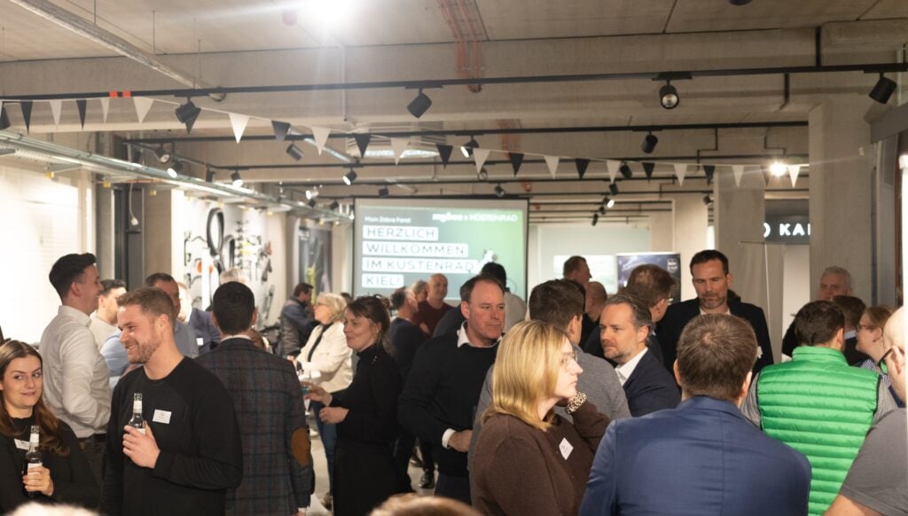 kuestenrad_Titel Eine Gruppe von Menschen unterhält sich bei einer Veranstaltung in einer Halle mit Bannern und einem Bildschirm mit der Aufschrift