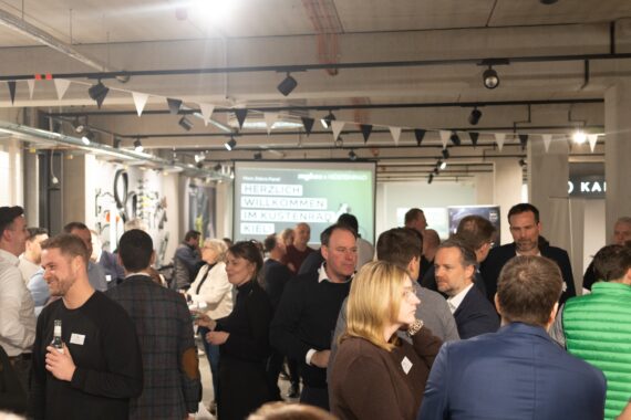 Eine Gruppe von Menschen unterhält sich bei einer Veranstaltung in einer Halle mit Bannern und einem Bildschirm mit der Aufschrift "HERZLICH WILLKOMMEN IM KÜSTENRAD KIE" auf Deutsch. Die Umgebung ist modern, mit Dekorationen und einigen Personen mit Namensschildern.