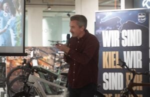 Ein Mann mit grauem Haar spricht bei einer Hallenveranstaltung neben Fahrrädern in ein Mikrofon. Hinter ihm steht ein großes Schild mit der Aufschrift WIR SIND KIEL WIR SIND und ein Teil eines projizierten Sportbildes.