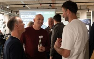 Eine Gruppe von Männern steht in einem Innenraum zusammen, lächelt und unterhält sich bei einem gesellschaftlichen Ereignis. Ein Mann hält ein Getränk in der Hand. Auf einem Schild im Hintergrund steht "HERZLICH WILLKOMMEN". Die Atmosphäre ist freundlich und locker.