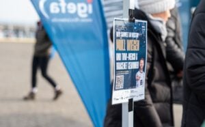 Ein Plakat ist an einem Pfahl im Freien befestigt, an dem unscharfe Menschen vorbeilaufen. Im Hintergrund sind blaue Banner mit dem Wort igefa zu sehen. Das Plakat enthält Text, Bilder und einen QR-Code.