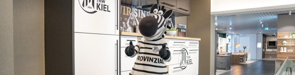 2526_Titel_Event_Küchen-Aktuell_1 Eine Person in einem Zebra-Maskottchen-Kostüm steht in einem modernen Büro oder Ausstellungsraum mit weißen Schränken, THW Kiel-Branding und Holzfußboden.