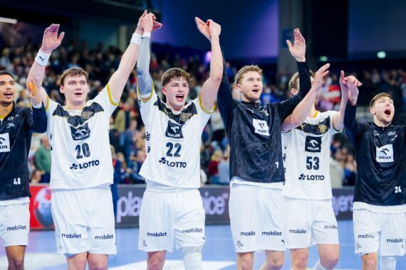 Fünf männliche Handballspieler in weißen und schwarzen Trikots stehen in einer Reihe, heben die Arme und jubeln, während im Hintergrund eine jubelnde Menge und helle Stadionlichter zu sehen sind.
