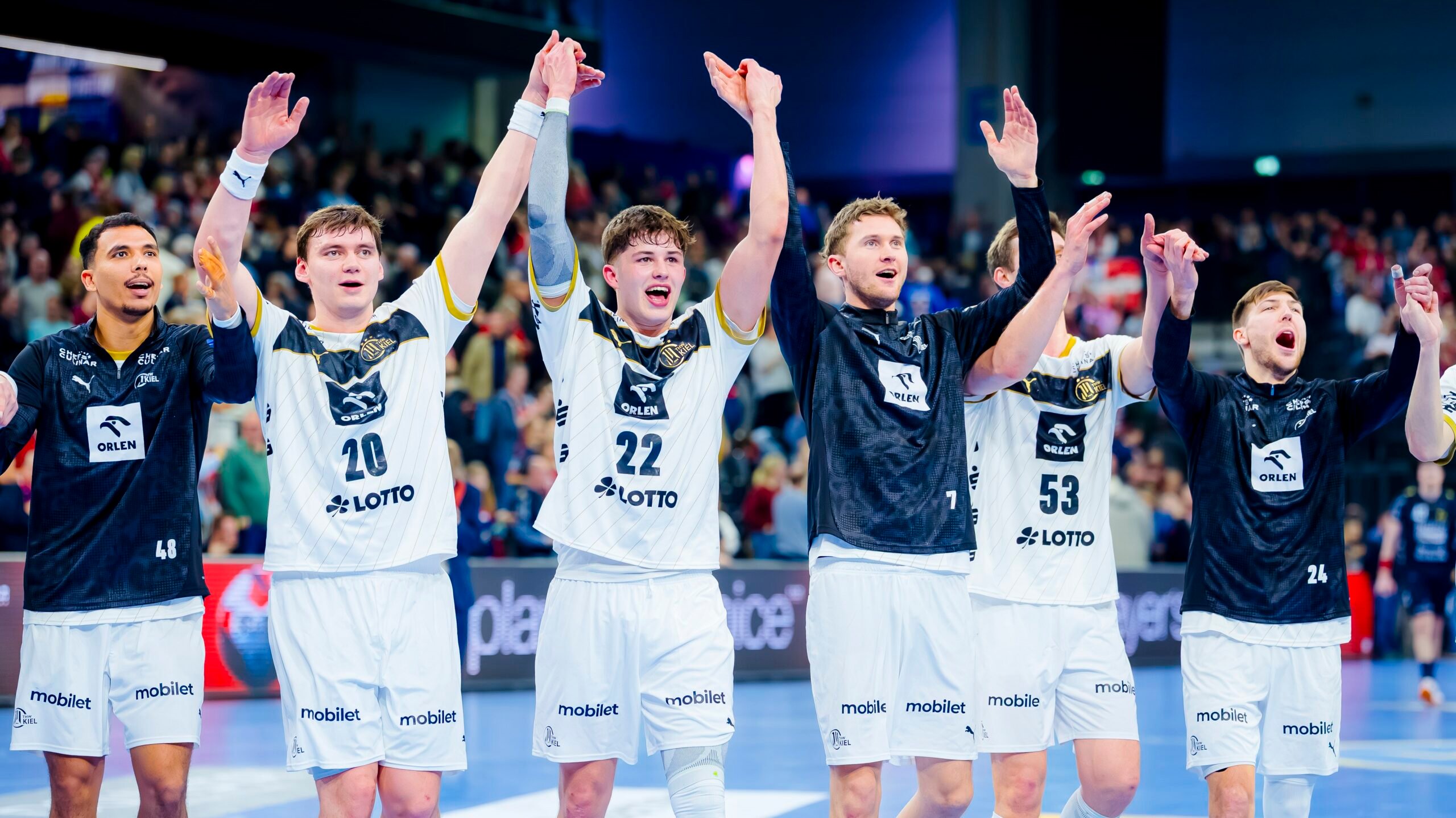 Fünf männliche Handballspieler in weißen und schwarzen Trikots stehen in einer Reihe, heben die Arme und jubeln, während im Hintergrund eine jubelnde Menge und helle Stadionlichter zu sehen sind.