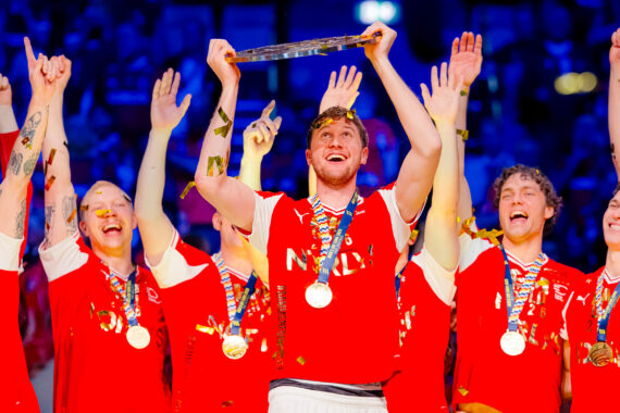 Ein jubelndes Männerhandballteam in roten Trikots, mit Medaillen um den Hals, feiert, während ein Spieler inmitten von Konfetti und jubelnden Mannschaftskameraden eine Trophäe über seinen Kopf hebt.