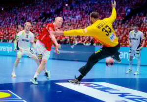 Ein Handballspieler in einem roten Trikot schießt den Ball in Richtung Tor, während der Torwart in einem gelben Trikot aufspringt, um den Ball zu blocken, während andere Spieler und eine Menschenmenge im Hintergrund zu sehen sind.