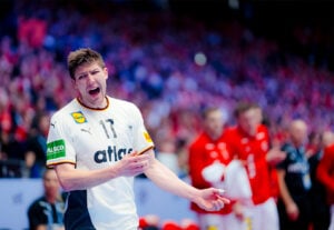 Ein männlicher Handballspieler in einem weißen Trikot mit der Nummer 17 reagiert mit Schmerzen und hält sich die Hand oder den Arm, während er auf einem Spielfeld steht. Im Hintergrund sind unscharfe Spieler in roten Trikots zu sehen.