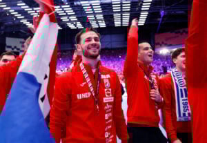 Eine Gruppe lächelnder Athleten in roten Jacken feiert in einer Halle, sie tragen Medaillen und winken, umgeben von einer jubelnden Menge und hellen Lichtern. Eine Person hält eine Fahne im Vordergrund.