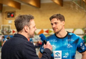 Ein Reporter mit einem blauen Mikrofon interviewt einen männlichen Sportler in einem blauen Sporttrikot in einer Sporthalle. Der Sportler schaut aufmerksam zu, während im Hintergrund unscharfe Sportgeräte und Menschen zu sehen sind.