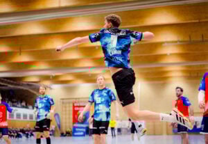 Ein männlicher Handballspieler in einem blauen Trikot springt in die Luft, um den Ball zu werfen, während andere Spieler in blauen und roten Trikots auf einem Hallenplatz zusehen.