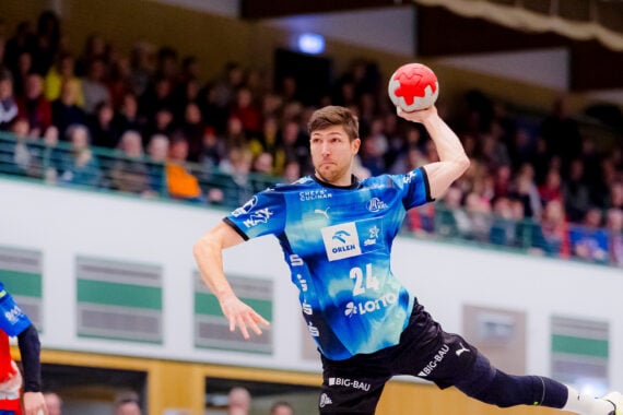 Ein männlicher Handballspieler in einem blauen Trikot springt, um einen roten Ball während eines Spiels zu werfen, während im Hintergrund eine Zuschauermenge zusieht. Ein anderer Spieler in einem roten Trikot steht links.