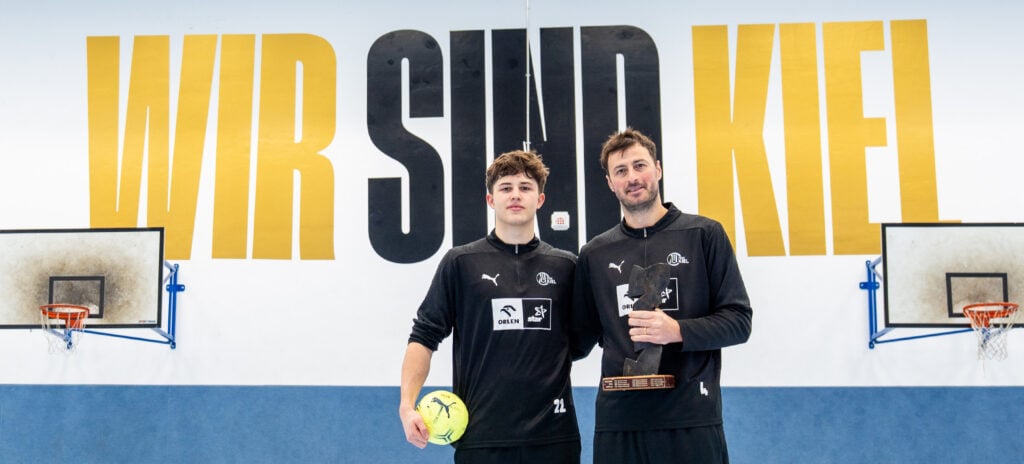 Zwei Männer in schwarzer Sportkleidung stehen in einer Turnhalle und halten einen gelben Fußball und einen Pokal in der Hand. Hinter ihnen steht in großen gelben Buchstaben WIR SIND KIEL an der Wand. Auf beiden Seiten sind Basketballkörbe zu sehen.