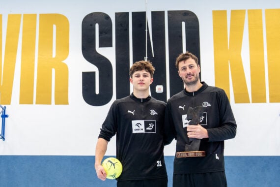 Zwei Männer in schwarzer Sportkleidung stehen in einer Turnhalle und halten einen gelben Fußball und einen Pokal in der Hand. Hinter ihnen steht in großen gelben Buchstaben WIR SIND KIEL an der Wand. Auf beiden Seiten sind Basketballkörbe zu sehen.