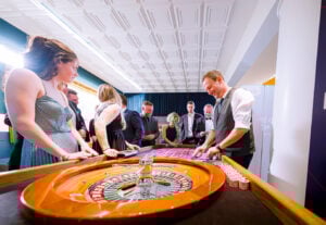 Eine Gruppe gut gekleideter Menschen versammelt sich um einen Roulettetisch im Kasino und beobachtet, wie der Croupier in einem hell erleuchteten, eleganten Raum das Rad dreht.