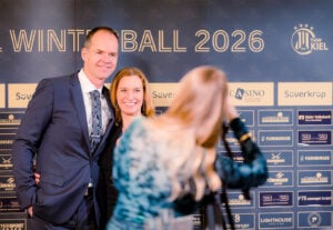 Ein Mann im Anzug und eine Frau im Abendkleid posieren zusammen und lächeln für ein Foto auf dem roten Teppich des Winterballs 2026, während im Vordergrund ein Fotograf ihr Bild aufnimmt.