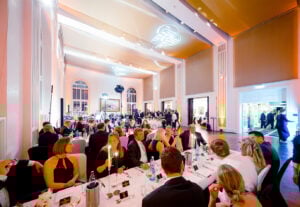 Ein großer, eleganter Bankettsaal, gefüllt mit gut gekleideten Gästen, die an runden Tischen sitzen und ein formelles Abendessen mit heller Beleuchtung und hohen Decken genießen.