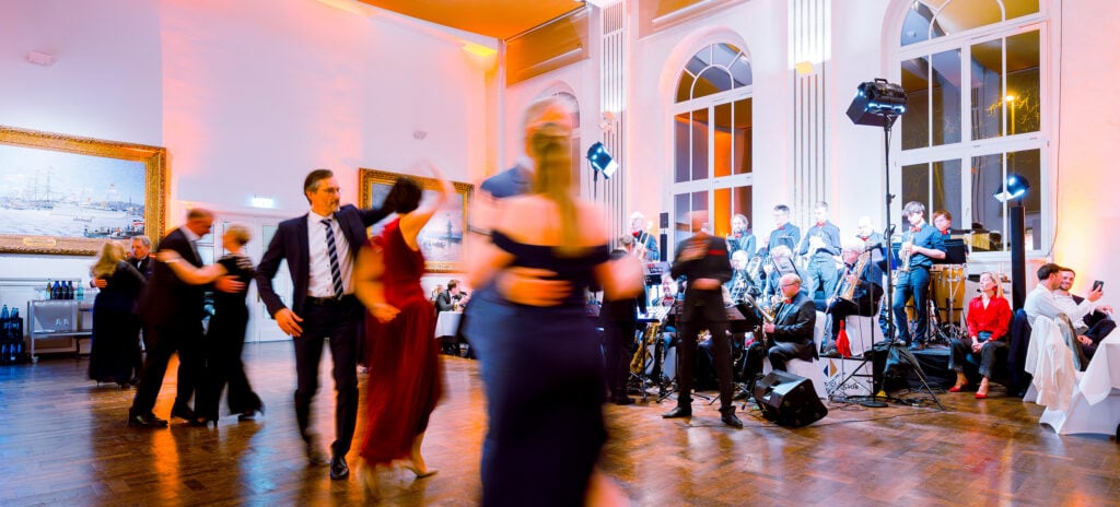 Menschen in formeller Kleidung tanzen in einem hell erleuchteten Ballsaal, während auf der Bühne eine Live-Band spielt. Große Fenster, Gemälde und elegante Dekoration sind im Hintergrund zu sehen. Die Szene wirkt lebendig und festlich.