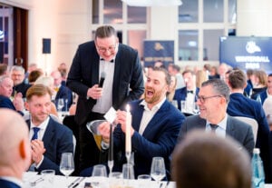 Ein gut gekleideter Mann steht mit einem Mikrofon und unterhält sich mit lachenden Gästen, die bei einem formellen Abendessen sitzen. Die lebhafte Menge, die geschmückten Tische und die elegante Atmosphäre lassen auf eine festliche Feier oder Gala schließen.