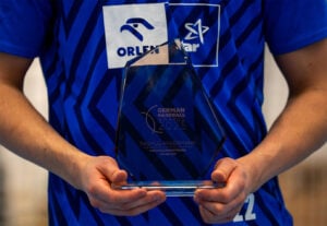 Eine Person in einem blauen Sporttrikot hält eine gläserne Trophäe für den Deutschen Handballpreis 2025, der an Rasmus Ankemann verliehen wurde. Auf dem Trikot sind Sponsorenlogos zu sehen, und die Hände der Person halten den Sockel der Trophäe.