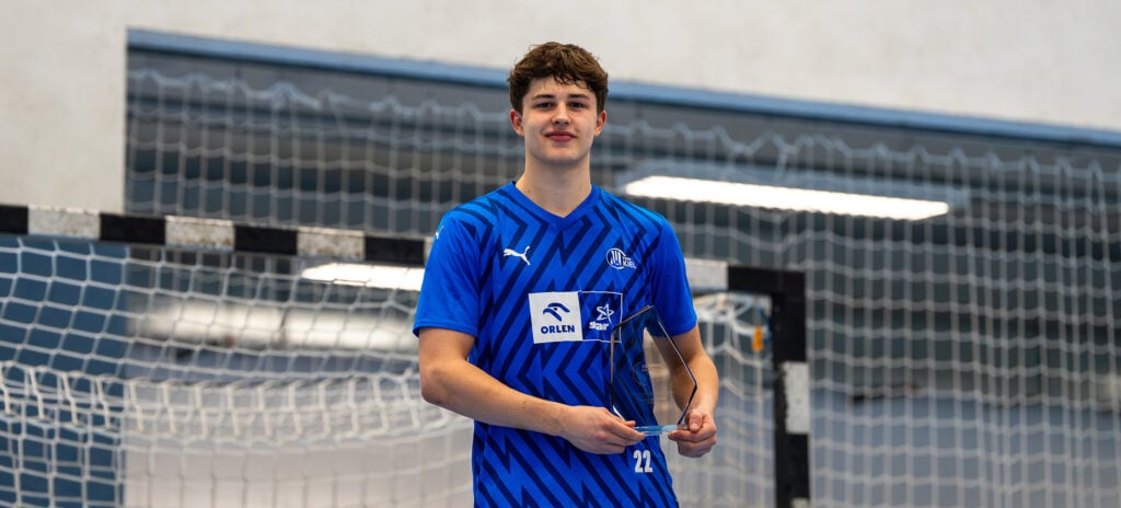 Ein junger männlicher Sportler in einem blauen Sportdress steht in einem Innenraum vor einem Handballtor, hält eine durchsichtige Trophäe in der Hand und lächelt in die Kamera.