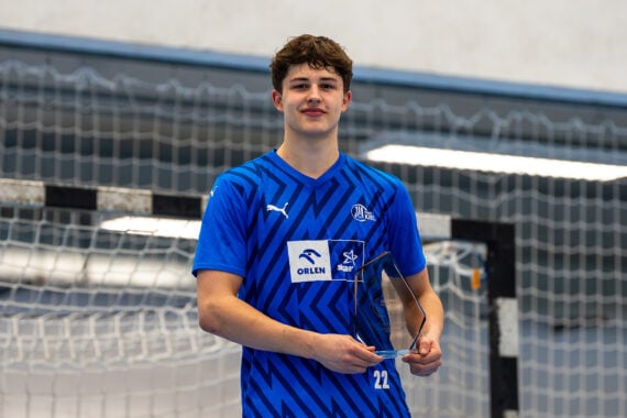 Ein junger männlicher Sportler in einem blauen Sportdress steht in einem Innenraum vor einem Handballtor, hält eine durchsichtige Trophäe in der Hand und lächelt in die Kamera.