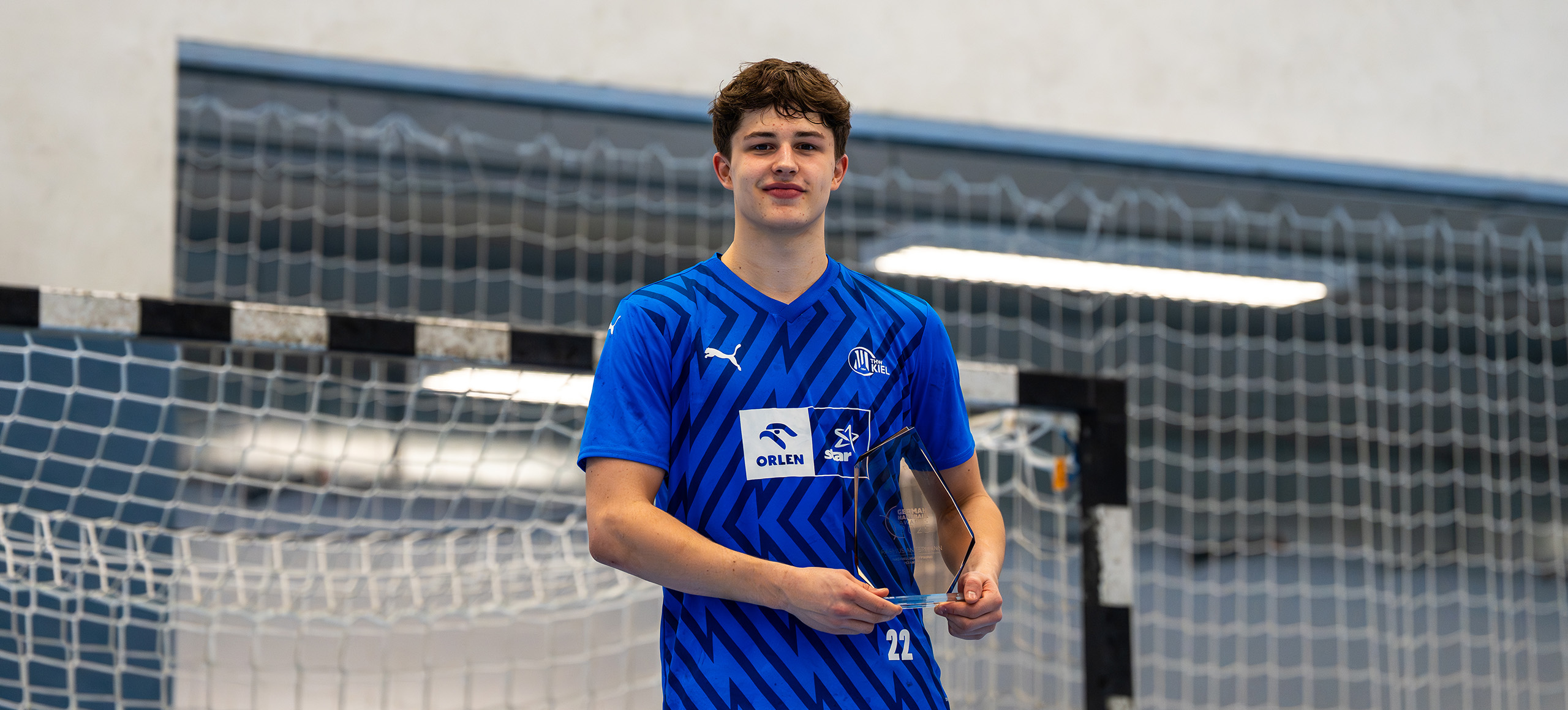 Ein junger männlicher Sportler in einem blauen Sportdress steht in einem Innenraum vor einem Handballtor, hält eine durchsichtige Trophäe in der Hand und lächelt in die Kamera.