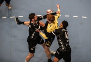 Zwei Handballspieler in schwarzen Trikots blockieren einen Spieler in einem gelben Trikot, der den Ball in der Luft hält, auf einem blauen Spielfeld. Die Szene ist intensiv und zeigt Körperkontakt und Action in der Nähe des Torraums.