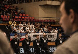 Eine Herren-Sportmannschaft steht in einer Reihe auf einem Spielfeld, mit Blick nach vorne, mit Fans im Hintergrund, die schwarz-weiß gekleidet sind und ein Wir sind Kiel-Banner halten. Rote und blaue Sitze füllen die Tribüne hinter ihnen.