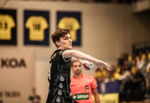 Ein männlicher Handballspieler in einem schwarzen Trikot bereitet sich darauf vor, den Ball während eines Spiels zu werfen. Der Hintergrund zeigt gelbe Trikots, die an der Wand hängen, und unscharfe Zuschauer auf der Tribüne.