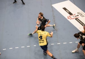 Ein Handballspieler in einem schwarzen Trikot läuft mit dem Ball, während ein Verteidiger in einem gelben Trikot versucht, ihn auf einem Hallenplatz zu blockieren. Andere Spieler und Spielfeldmarkierungen sind sichtbar.