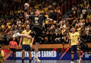 Ein Handballspieler in einem schwarzen Trikot springt in die Luft, um den Ball zu werfen, während die gegnerischen Spieler in gelben Trikots zusehen. Das Publikum füllt die Tribüne hinter ihnen, und auf der LED-Anzeige am Spielfeldrand ist ein fetter Text zu sehen.