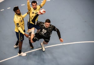 Ein Handballspieler in einem schwarzen Trikot versucht einen Sprungwurf, während er von zwei Spielern in gelben Trikots auf einem Hallenplatz eng verteidigt wird.