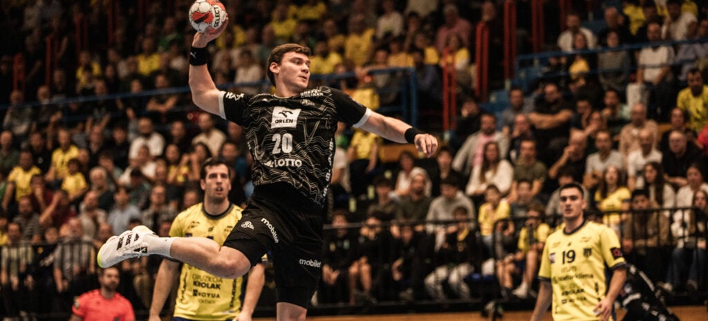 260224_Bidasoa_THW_Titel_Laube Ein Handballspieler in einem schwarzen Trikot springt in der Luft, um den Ball zu werfen, umgeben von gegnerischen Spielern in Gelb, während im Hintergrund eine große Menschenmenge von der Tribüne aus zusieht.