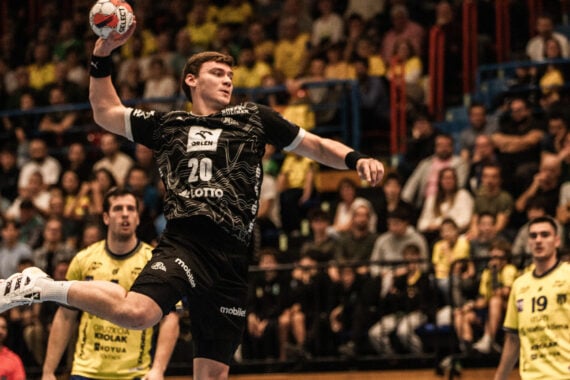 Ein Handballspieler in einem schwarzen Trikot springt in der Luft, um den Ball zu werfen, umgeben von gegnerischen Spielern in Gelb, während im Hintergrund eine große Menschenmenge von der Tribüne aus zusieht.