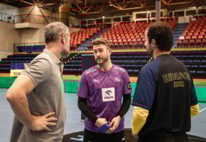 Drei Männer stehen auf einem überdachten Sportplatz und unterhalten sich. Einer trägt ein lila Trikot mit der Aufschrift "ORLEN", ein anderer ein schwarz-gelbes "BIDASOA IRUN"-Shirt und der dritte ein graues Polo. Im Hintergrund sind leere rote und blaue Sitzplätze zu sehen.
