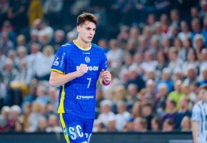 Ein Handballspieler in einem blau-gelben Trikot mit der Nummer 7 joggt auf einem Hallenplatz, während im Hintergrund eine große, unscharfe Zuschauermenge zu sehen ist.