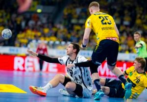 Ein männlicher Handballspieler in einem weißen Trikot sitzt auf dem Spielfeld und greift nach dem Ball, während zwei gegnerische Spieler in gelben Trikots während eines intensiven Spiels in einer Halle daneben stehen und knien.