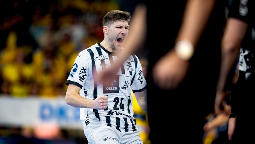 Ein männlicher Handballspieler in einem weiß-schwarzen Trikot jubelt leidenschaftlich auf dem Spielfeld, ballt die Faust und schreit, während im Hintergrund verschwommene Figuren und Zuschauer zu sehen sind.