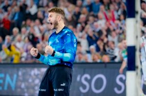 Ein männlicher Sportler in einem blauen Trikot feiert leidenschaftlich mit geballten Fäusten und offenem Mund vor einer verschwommenen Menge in einer Sporthalle.